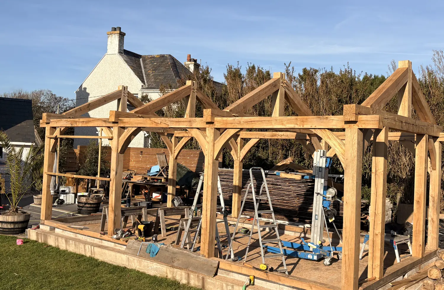 A green oak frame raised on its base, posts, braces and four trusses standing against blue sky with the house behind.