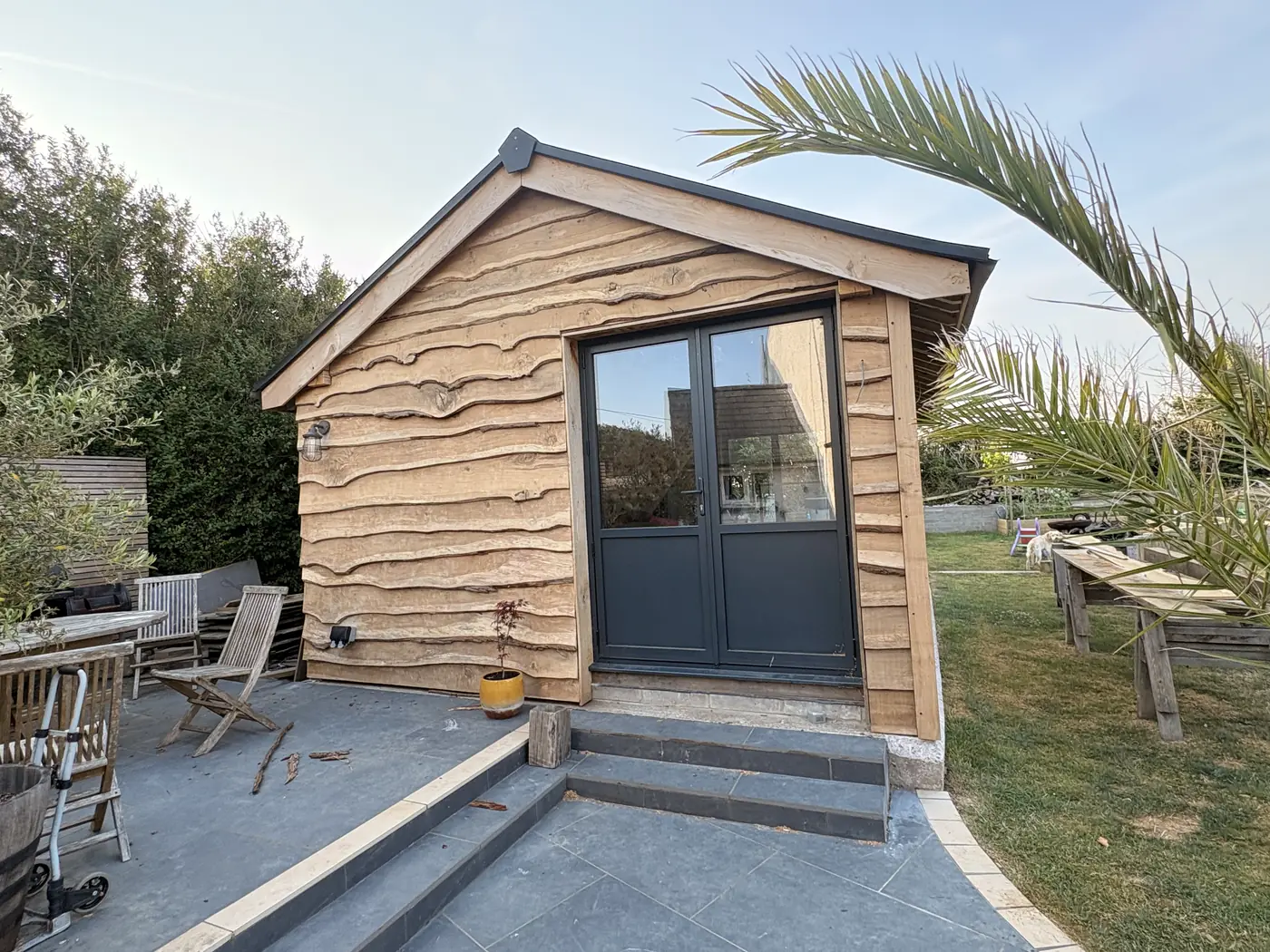 A finished oak framed garden room clad in waney-edge oak, with dark glazed doors, on a paved terrace.