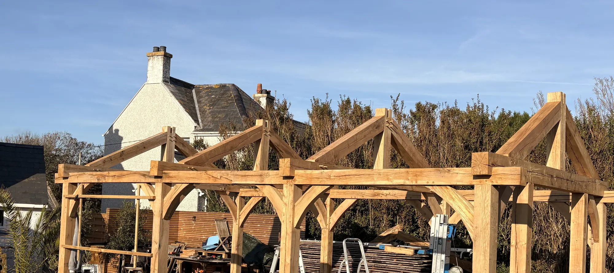 Four green oak trusses with curved braces, standing raised against clear blue sky, the house behind.
