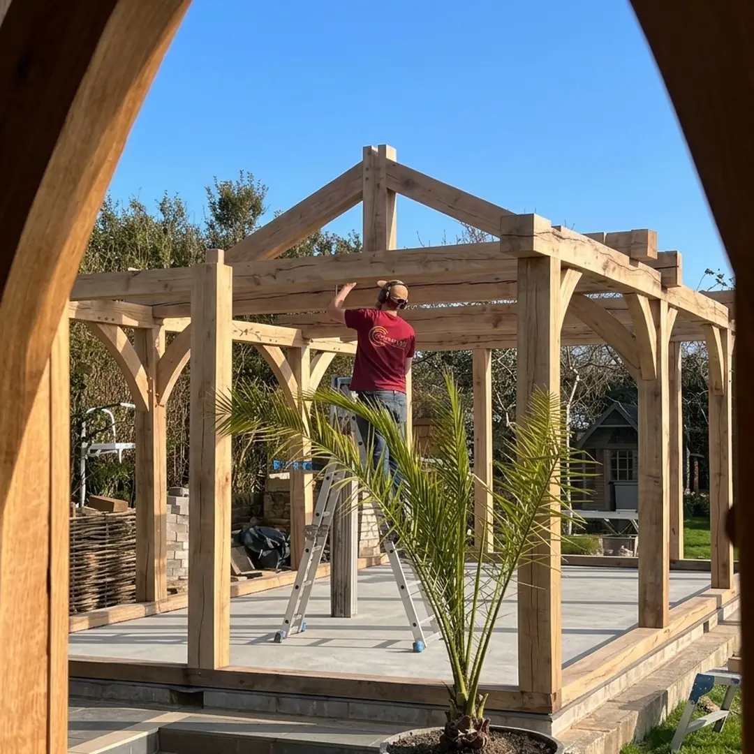 A carpenter on a step ladder fitting a tie beam into a green oak frame being raised on site.