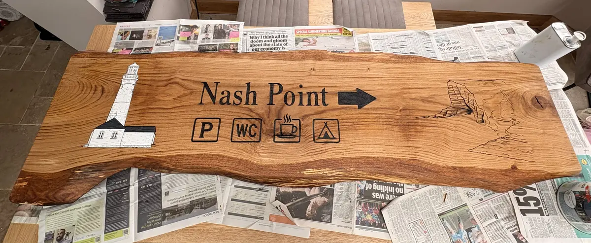A long live-edge oak board engraved with the words Nash Point, a lighthouse illustration, a directional arrow and parking, toilet and picnic symbols.